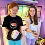 Swiss Cows Children holding a small pet and a snake in a room with a reptile tank and the word 'SNAKE' on the wall.
