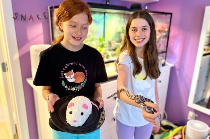 Children holding a small pet and a snake in a room with a reptile tank and the word 'SNAKE' on the wall.