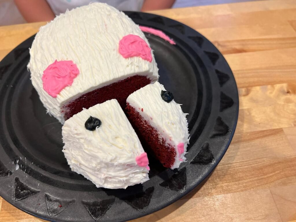 A cute pig-shaped red velvet cake with white frosting and pink accents served on a black decorative plate.