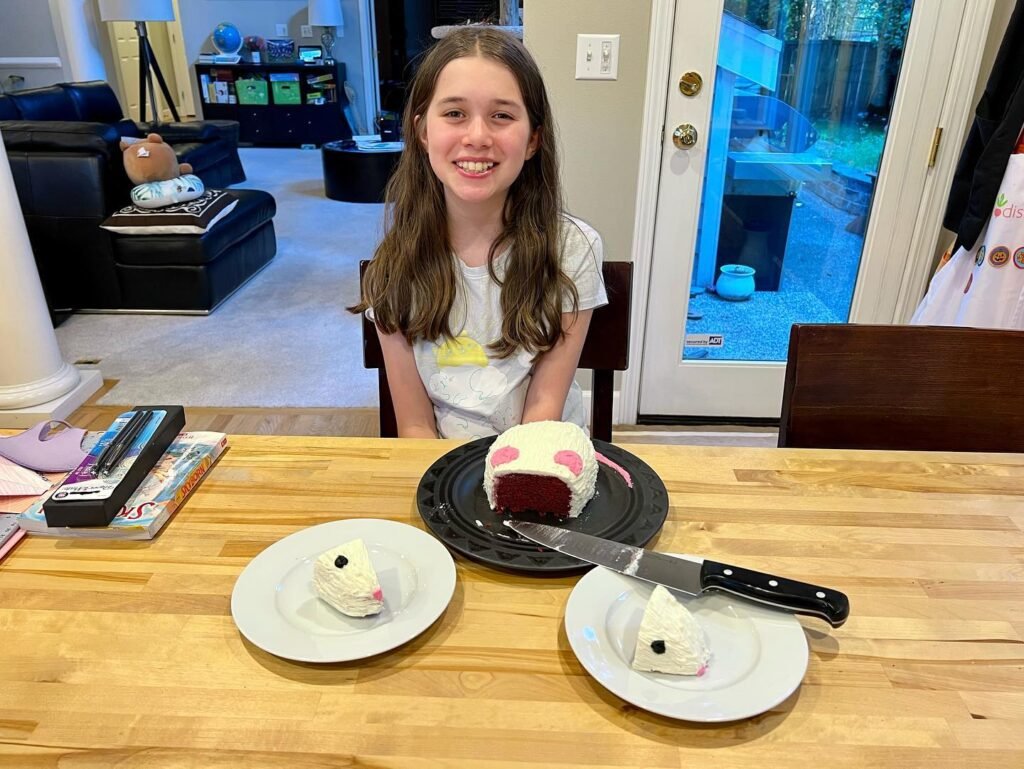 A smiling person is enjoying a slice of red velvet cake with frosted decorations on a wooden dining table. The cozy home environment adds warmth to the moment.