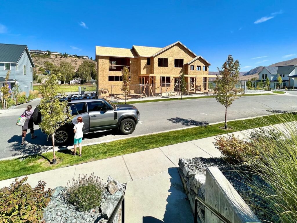 A two-story home under construction with a bare exterior frame on a sunny day. A pickup truck is parked on the quiet residential street.