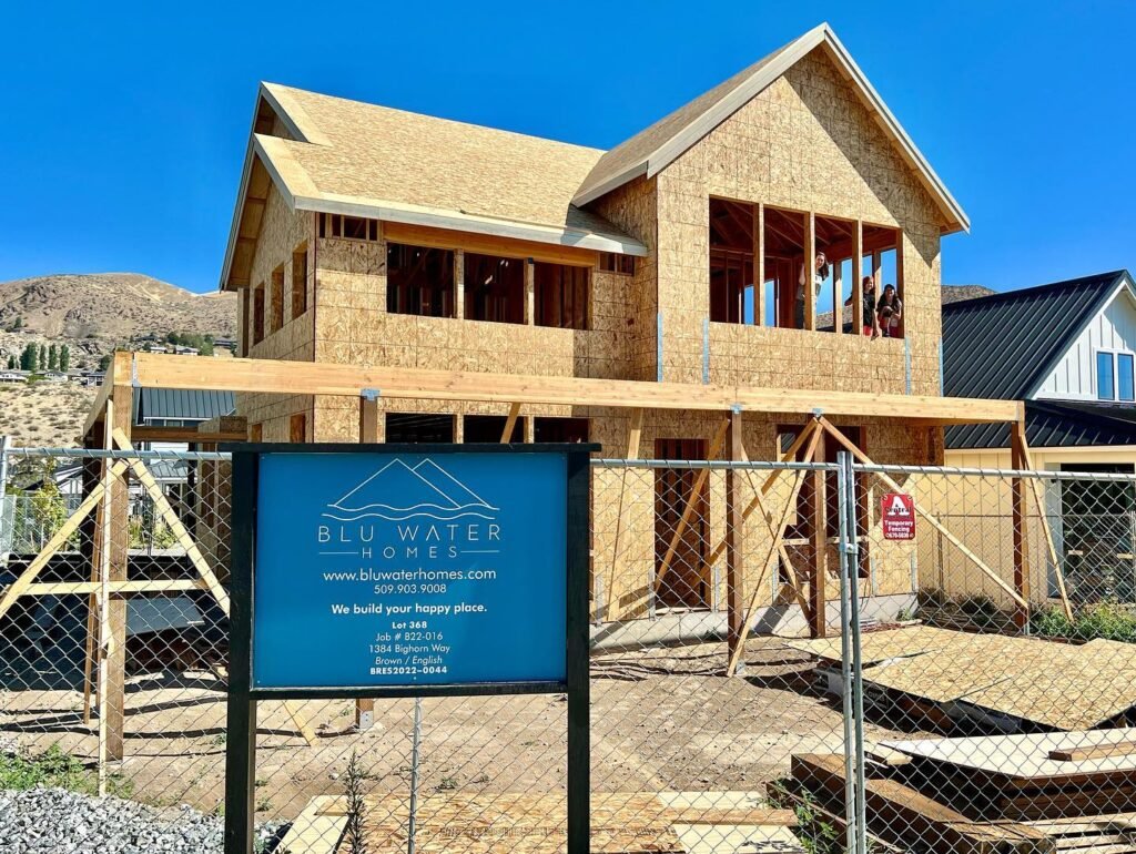 A new home under construction with a Blue Water Roofing sign prominently displayed, framed against hills and a blue sky.