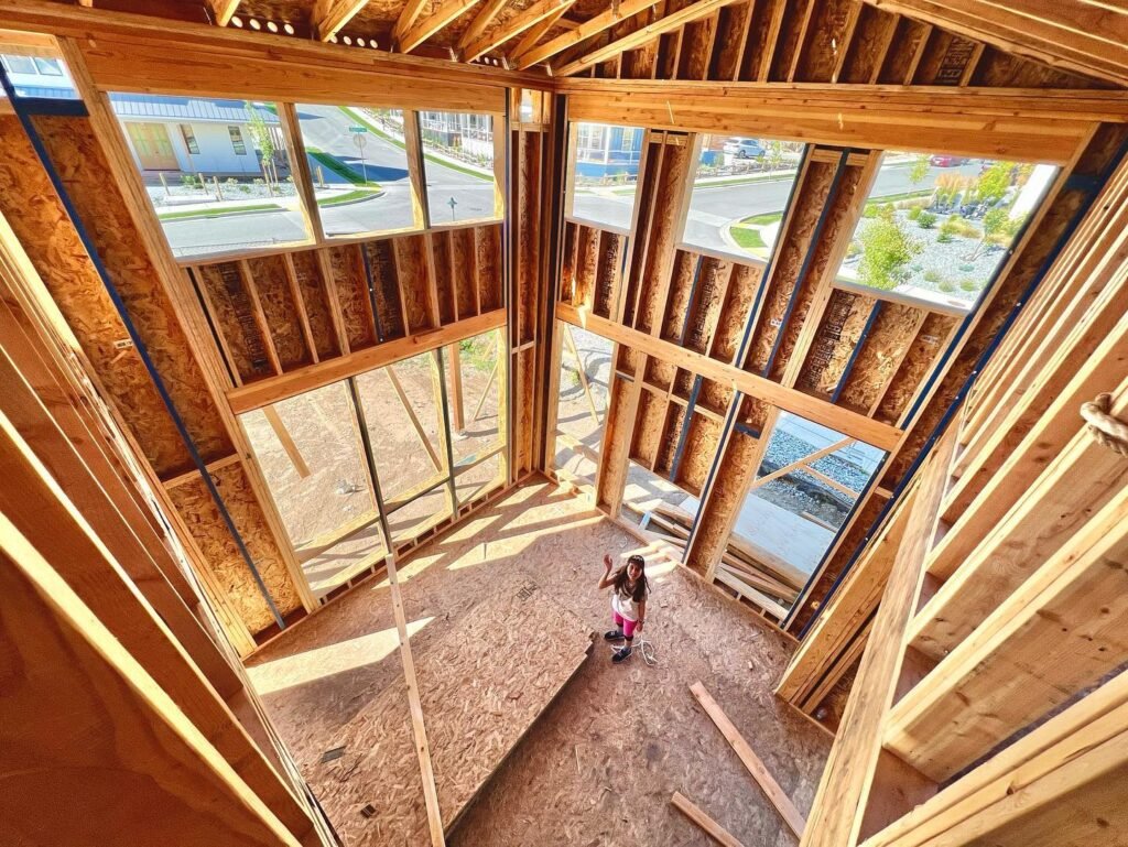 A view from inside an unfinished house showing wooden framing and large window openings facing a residential street.