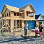 A view of our new home under construction on a sunny day. Two people stand in front of the framed structure with a blue sky and hills in the background.