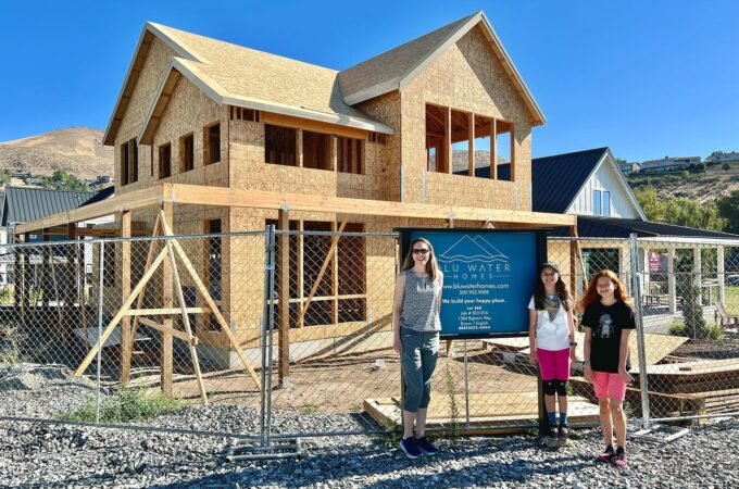 A view of our new home under construction on a sunny day. Two people stand in front of the framed structure with a blue sky and hills in the background.