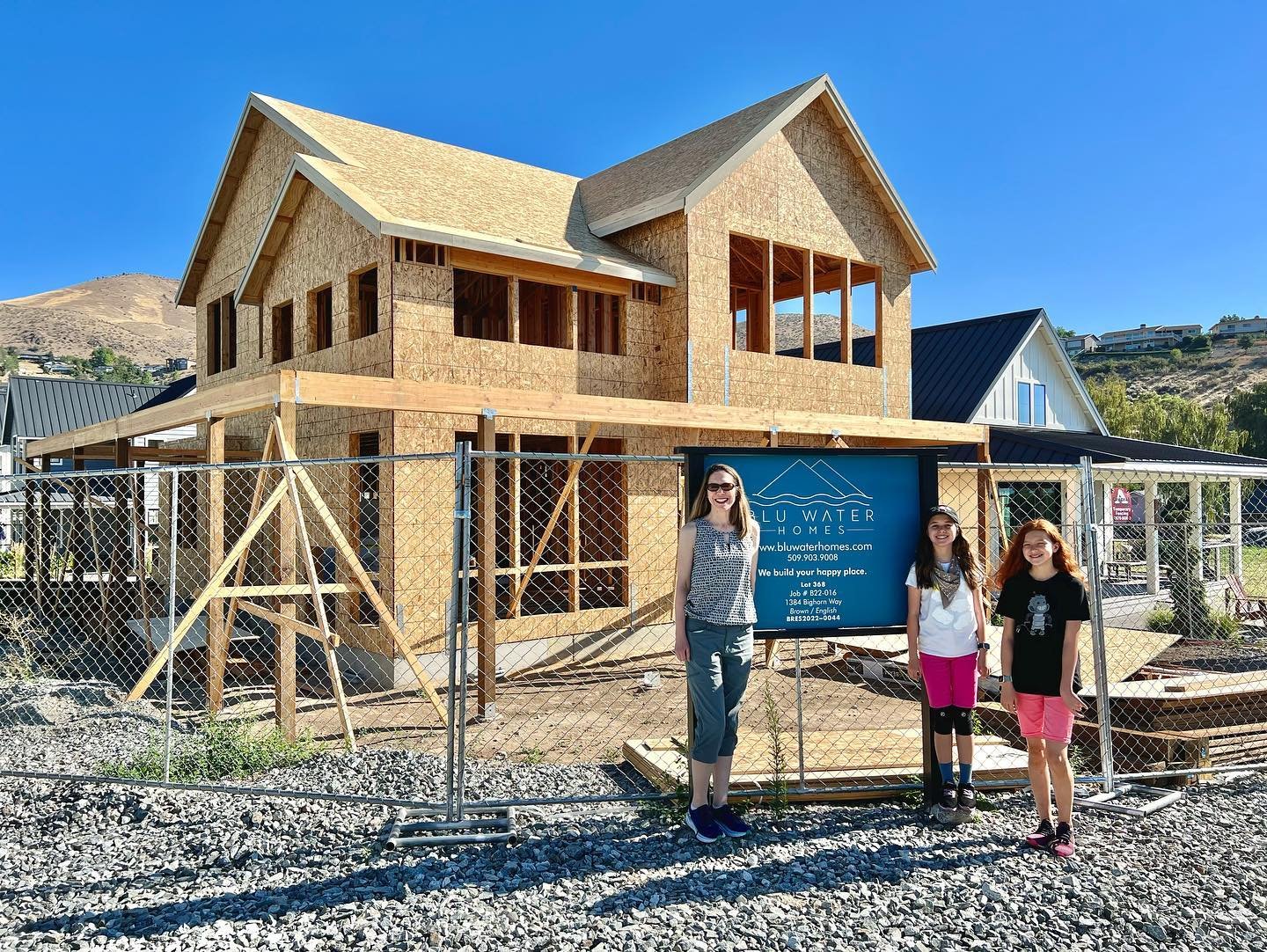 A view of our new home under construction on a sunny day. Two people stand in front of the framed structure with a blue sky and hills in the background.