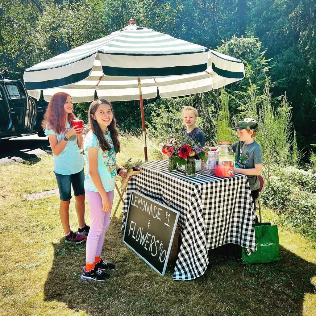 Outdoor lemonade stand with a striped umbrella and a checkered tablecloth. Group of people around the stand holding a drink.