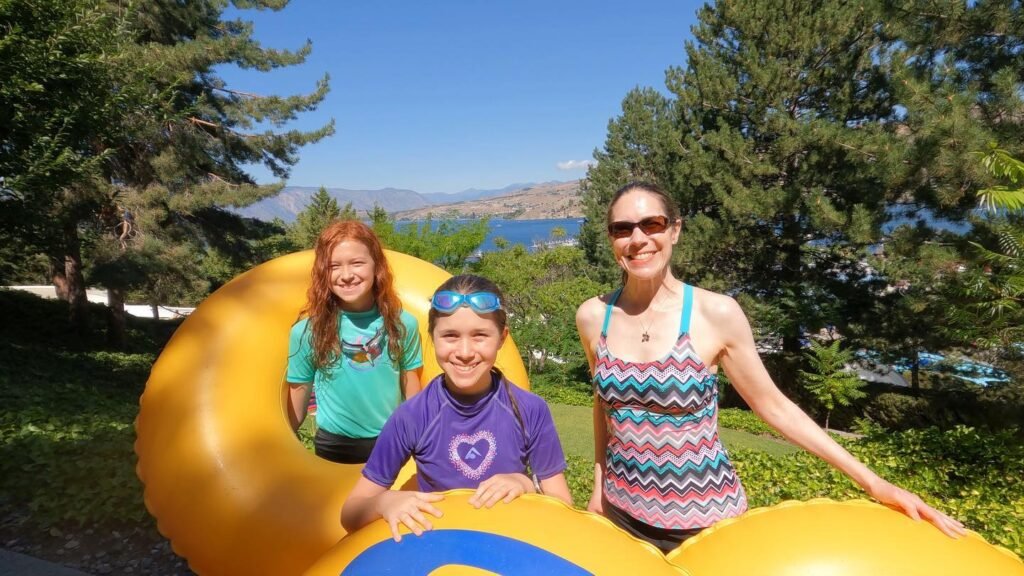 Three people enjoy a sunny day outdoors, posing near a large yellow inflatable structure with trees and water in the background.