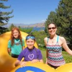 Three people enjoy a sunny day outdoors, posing near a large yellow inflatable structure with trees and water in the background.