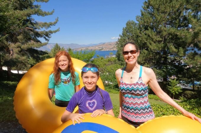Three people enjoy a sunny day outdoors, posing near a large yellow inflatable structure with trees and water in the background.