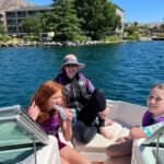 Chelanigans A group of three people wearing life jackets enjoys a boat ride on a calm lake with buildings in the background.