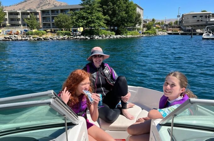 A group of three people wearing life jackets enjoys a boat ride on a calm lake with buildings in the background.