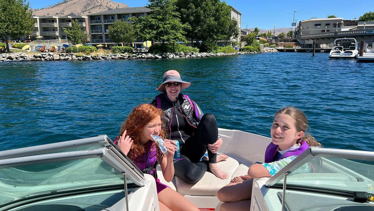 A group of three people wearing life jackets enjoys a boat ride on a calm lake with buildings in the background.