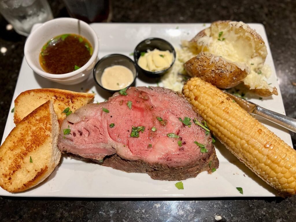 Close-up view of a plated filet mignon dinner featuring grilled bread, corn, potatoes, and dipping sauces.