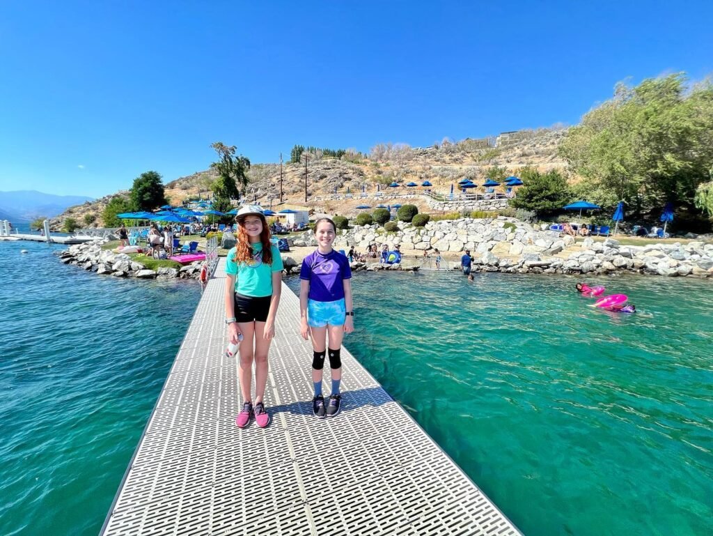 Two people stand on a metal pier extending into the turquoise waters of a mountain lake on a clear sunny day.