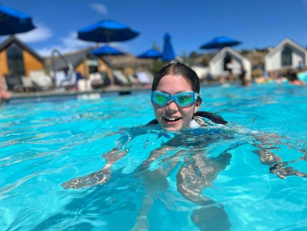 A person with a smile is swimming in vibrant resort pool water. In the background, colorful bungalows feature blue umbrellas and loungers.