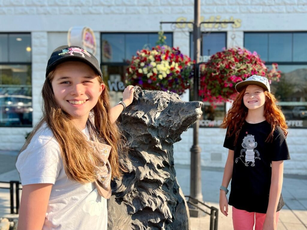 Two young girls pose together on a paved outdoor path beside a stone bear statue with hanging flower baskets in the background.