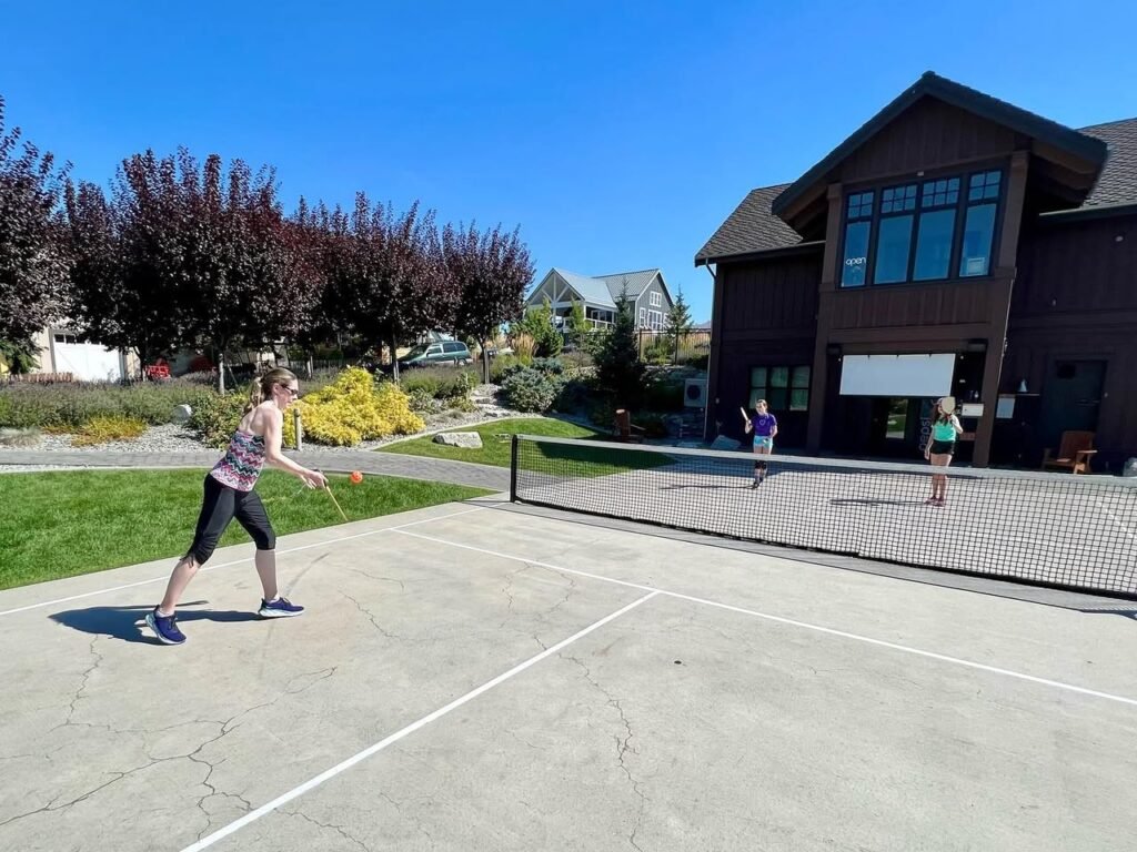 A group of people playing tennis on a sunny day in the Pacific Northwest. A dark wooden building and vibrant landscape are visible in the background.