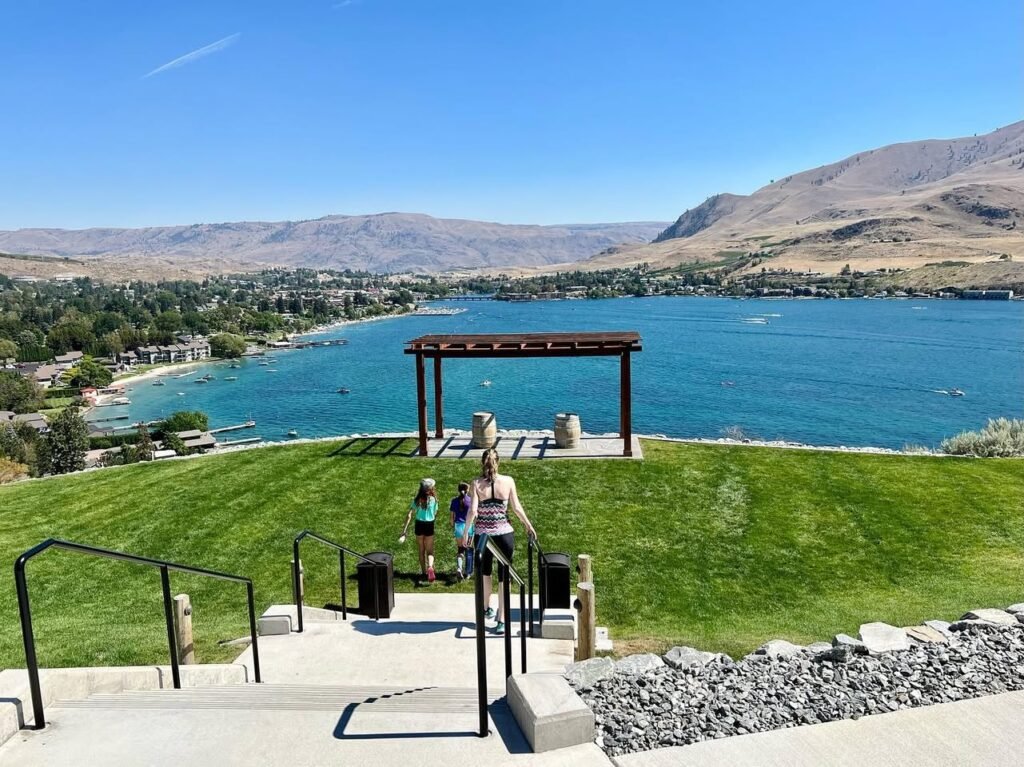 Concrete stairs with black railings leading down to a lake bordered by green lawns and mountains under a blue sky.