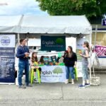 Chelanigans A company booth at an outdoor market features English Insurance Group and Bay Fair Fitness banners. Several people are gathered at the table under a white canopy tent.