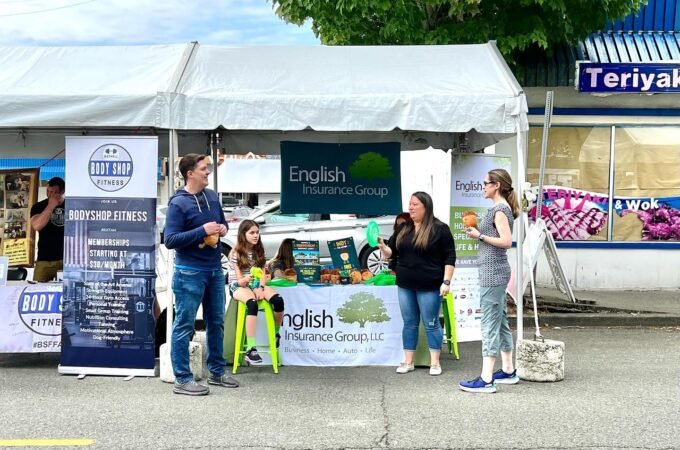 A company booth at an outdoor market features English Insurance Group and Bay Fair Fitness banners. Several people are gathered at the table under a white canopy tent.
