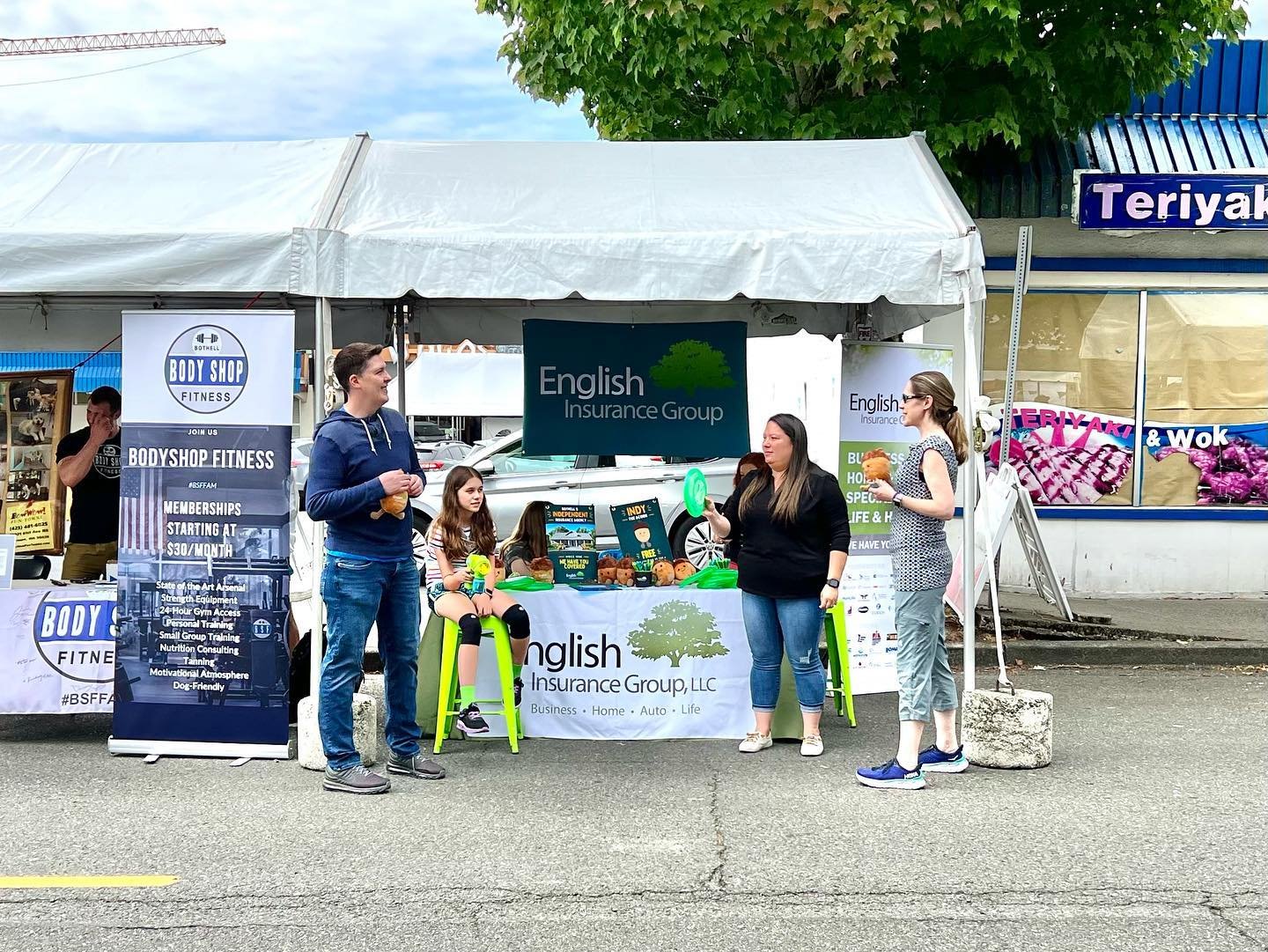 Bothell Summerfest A company booth at an outdoor market features English Insurance Group and Bay Fair Fitness banners. Several people are gathered at the table under a white canopy tent.