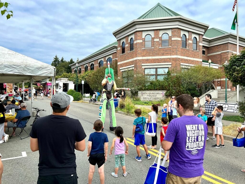 People watch a stilt walker perform during a street festival near a brick building with a green roof.