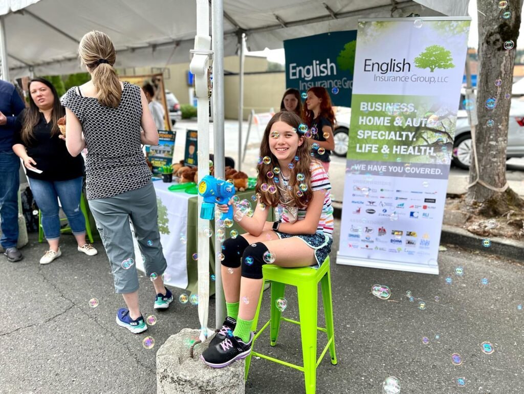 A youth enjoys a summer day at an outdoor event, seated on a green stool under a white tent. Banners and onlookers are visible in the background, with bubbles floating around.