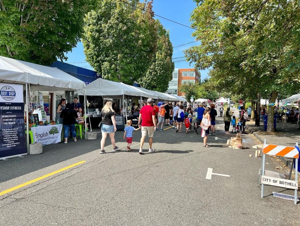 Crowded outdoor market with vendor tents lining a tree-lined street under a clear blue sky in the Pacific Northwest.