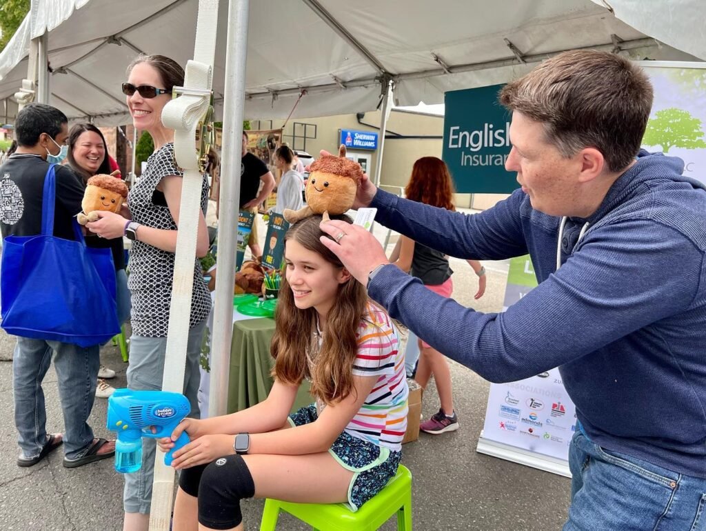 An adult playfully placing a toy squirrel on a child's head at a market tent. An English Insurance banner advertises the event in the background.