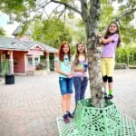 A group of individuals posing around a tree in a park, with a cast iron structure around its base. A unique pink building stands in the background.
