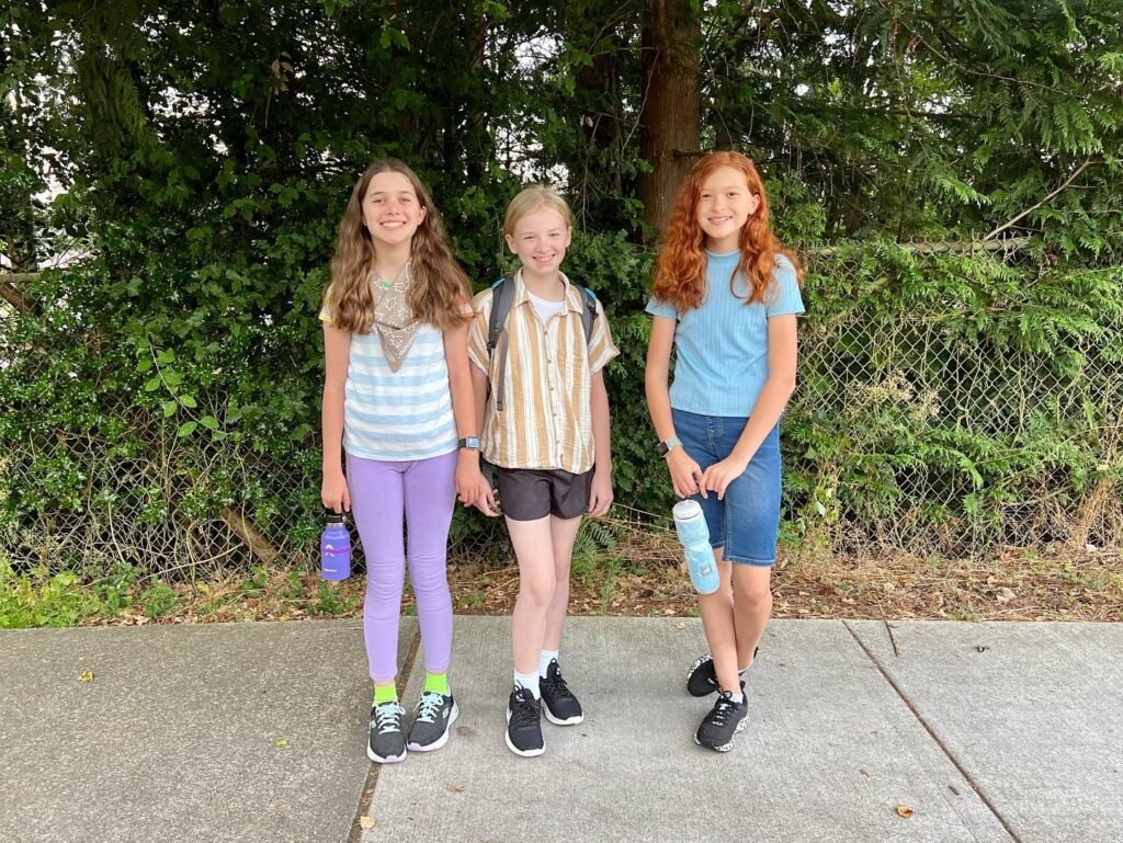 Three young girls holding hands and walking on a concrete path near evergreen trees.