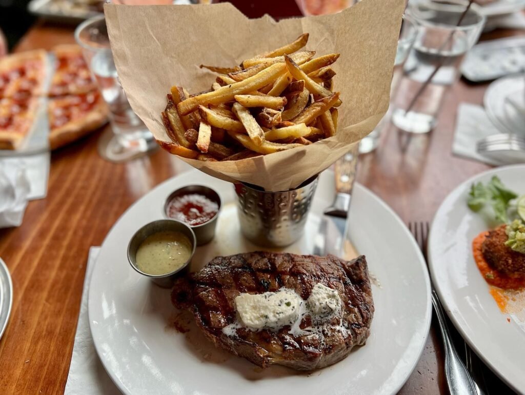 Grilled steak topped with herbs served with side dishes, including fries and dipping sauces, on a wooden dining table.