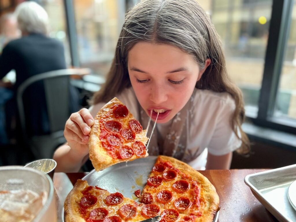 A young girl with long hair is happily eating a slice of pepperoni pizza. Sticky cheese stretches from the slice as she takes a bite in a cozy restaurant setting.