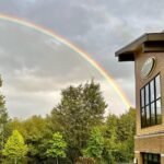 A striking rainbow arches gracefully across a cloudy sky above a line of lush green trees. A modern brick building with large windows stands prominently on the right side of the frame.