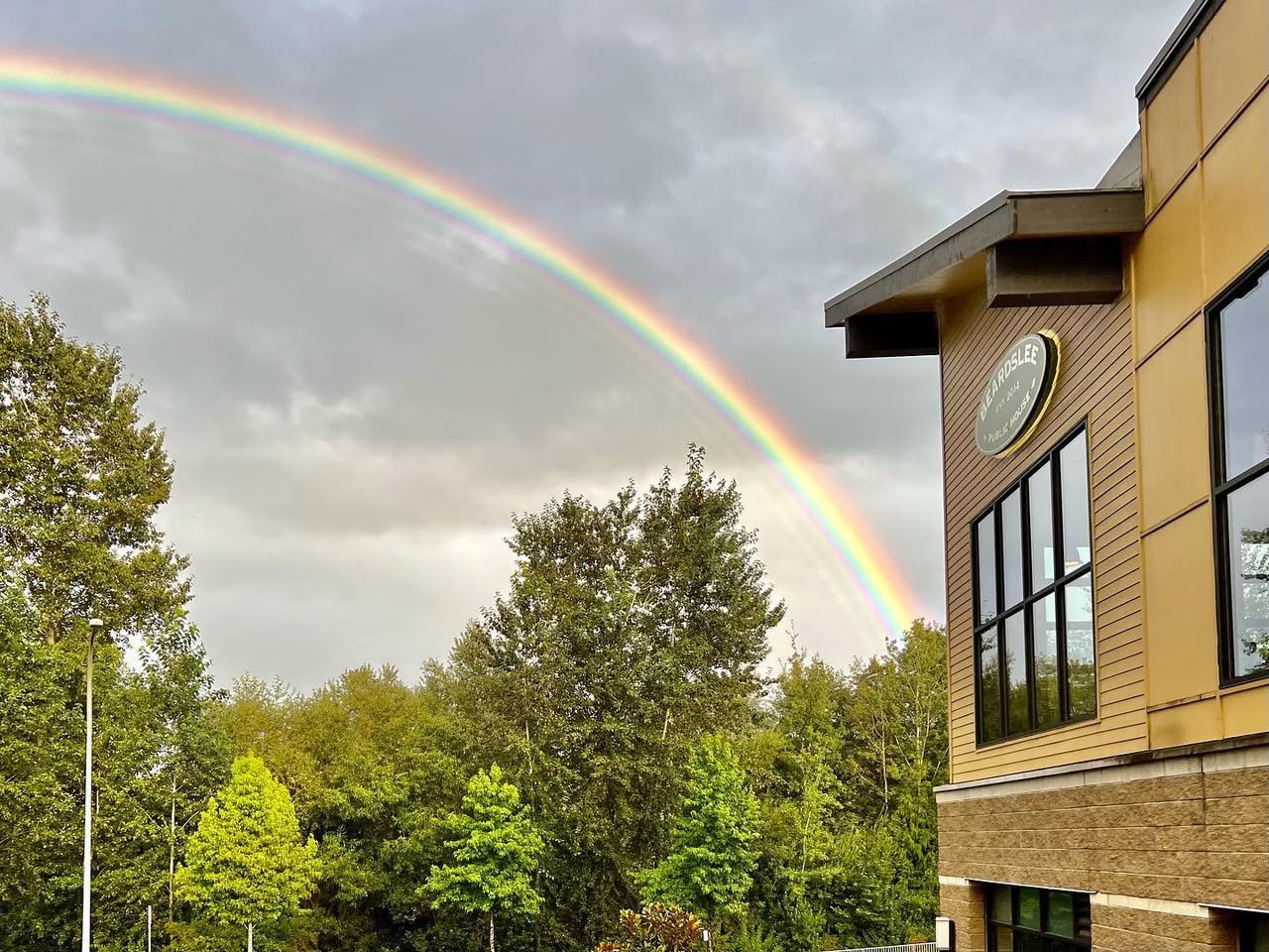 Beardslee Public House A striking rainbow arches gracefully across a cloudy sky above a line of lush green trees. A modern brick building with large windows stands prominently on the right side of the frame.