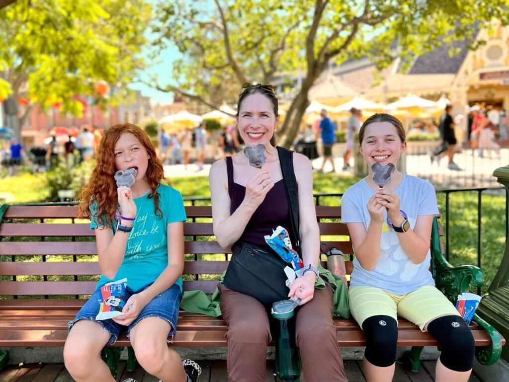 Three people sitting on a park bench enjoying soft-serve ice cream on a sunny day.