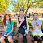 Three people sitting on a park bench enjoying soft-serve ice cream on a sunny day.