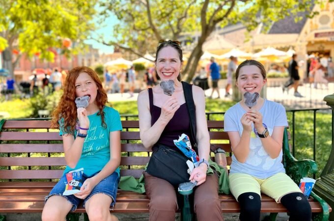 Three people sitting on a park bench enjoying soft-serve ice cream on a sunny day.