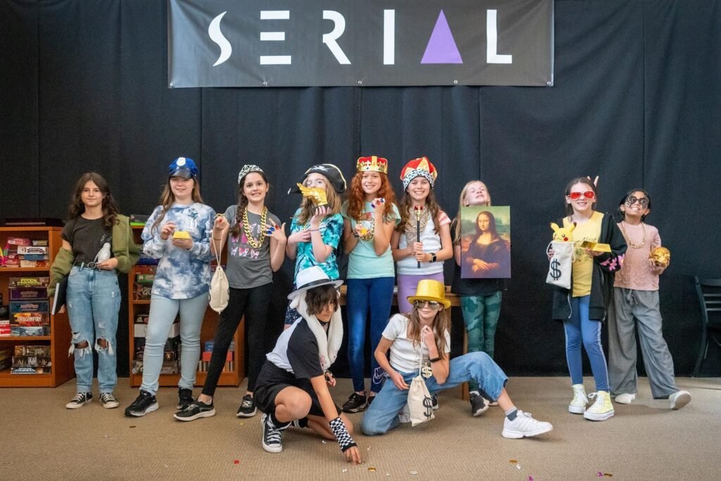 A diverse group of students in creative costumes poses in front of a black curtain with a 'SERIAL' banner and bookshelves.