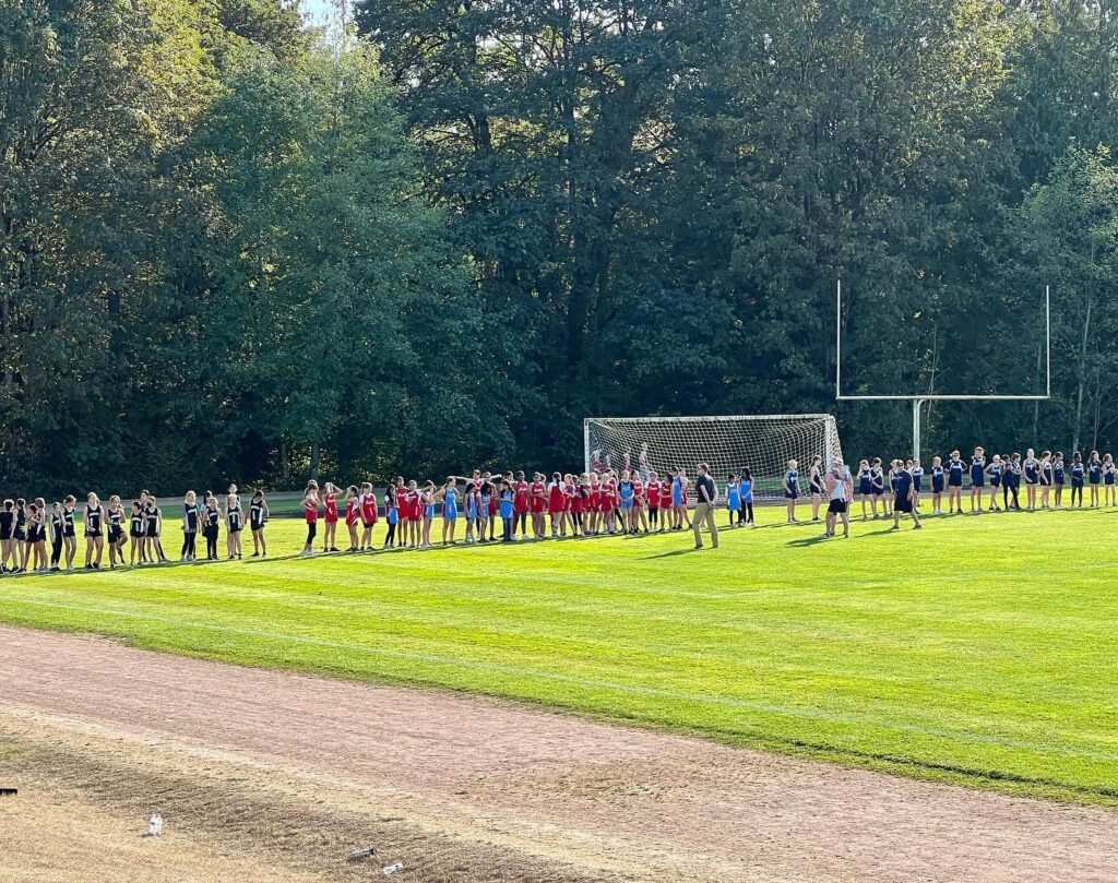 Groups of people in red and black uniforms stand on a green sports field near a running track with a goal net in the background.