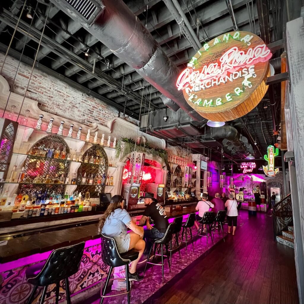 Interior of a colorful karaoke bar in Bangkok, featuring vibrant neon lights, a long wooden bar, and patrons seated on high stools.