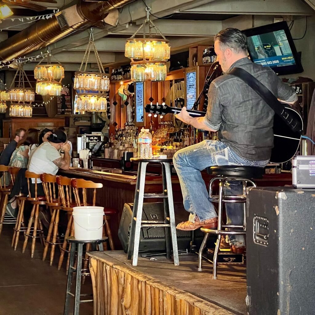 A musician plays guitar on a small stage in a bustling bar, surrounded by patrons and glowing lanterns.