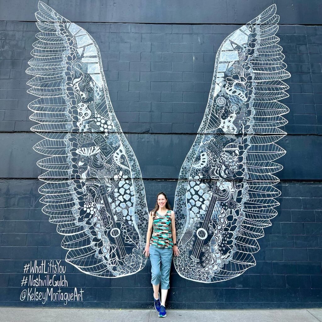 Detailed chalk artwork of angel wings on a dark wall in Nashville Gulch. A traveler poses beside the stunning public art.