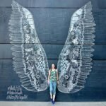 Detailed chalk artwork of angel wings on a dark wall in Nashville Gulch. A traveler poses beside the stunning public art.