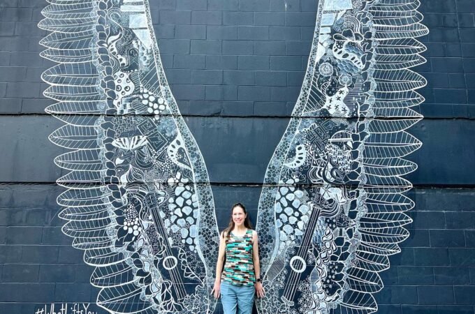 Detailed chalk artwork of angel wings on a dark wall in Nashville Gulch. A traveler poses beside the stunning public art.