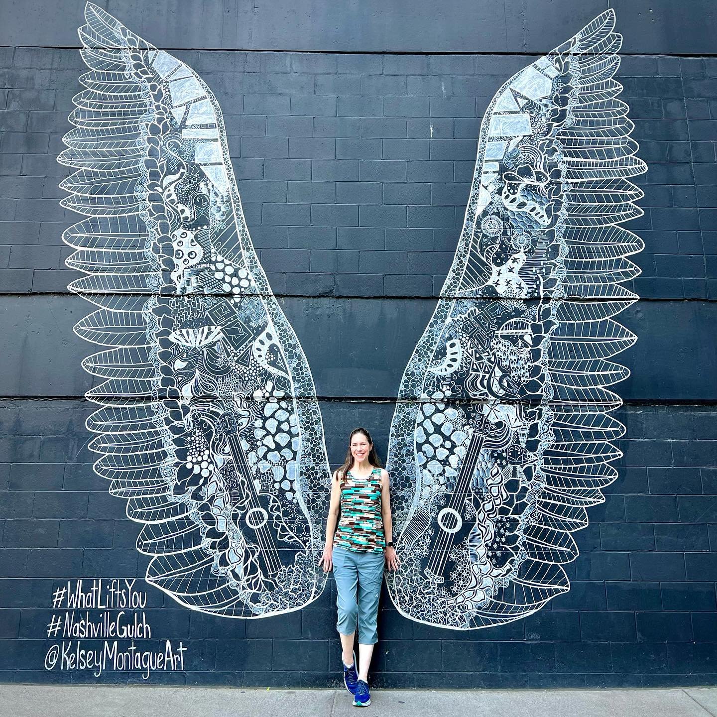 Detailed chalk artwork of angel wings on a dark wall in Nashville Gulch. A traveler poses beside the stunning public art.