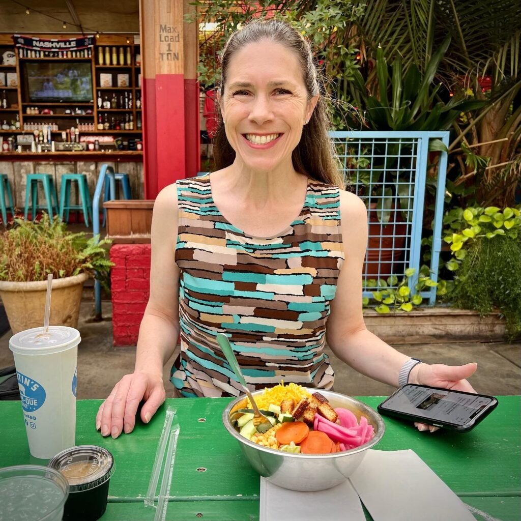 A smiling woman poses with a healthy smoothie bowl at a vibrant outdoor cafe. The setting includes lush tropical plants, a green table, and a casual bar area in the background.