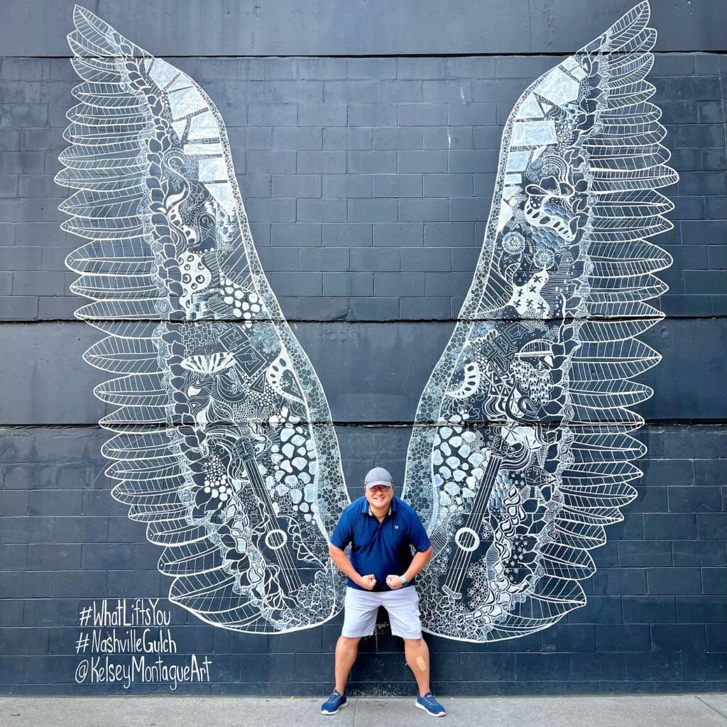 A person standing in front of a detailed mural of wings on a dark brick wall. The wings are composed of numerous small, intricate objects.