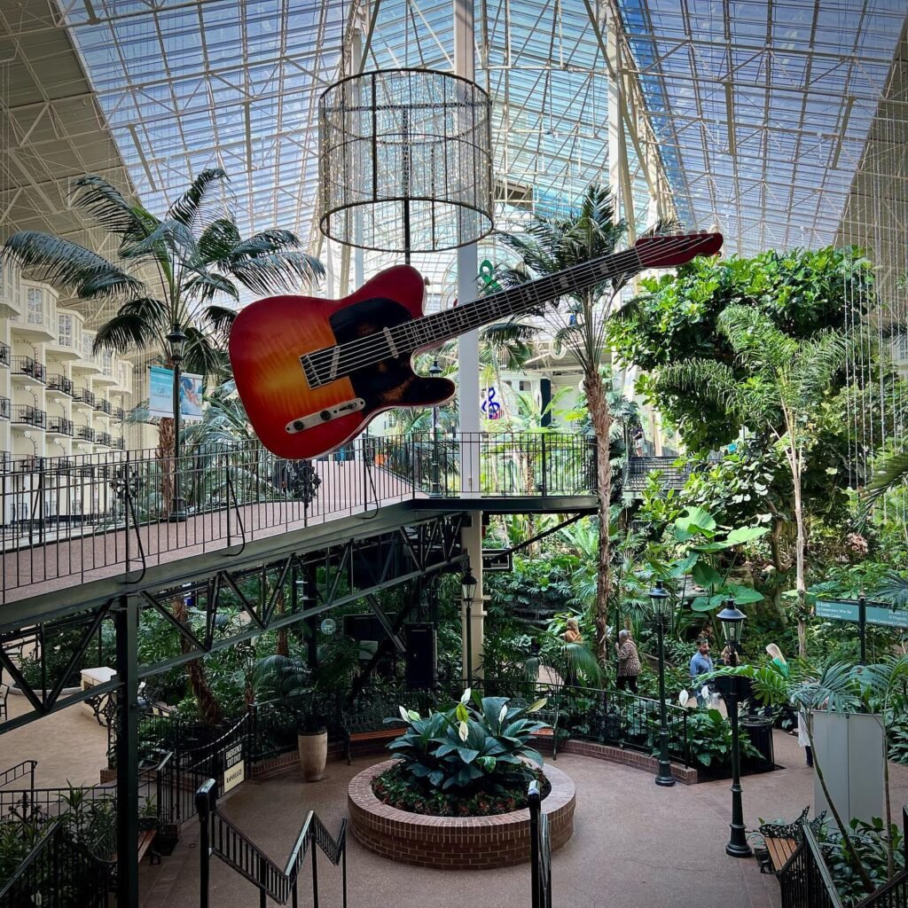 A vibrant electric guitar hangs suspended in a lush, glass-roofed conservatory filled with tropical plants and multi-level walkways.